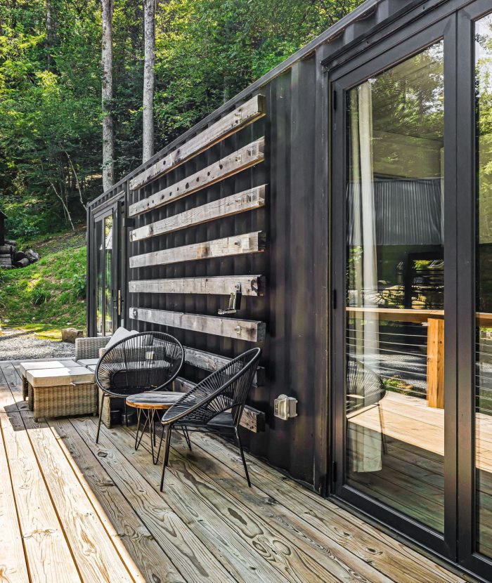 A modern cabin with a wooden deck, black exterior, and glass sliding doors. There are two chairs and a small table on the deck, set amidst a lush, forested background.