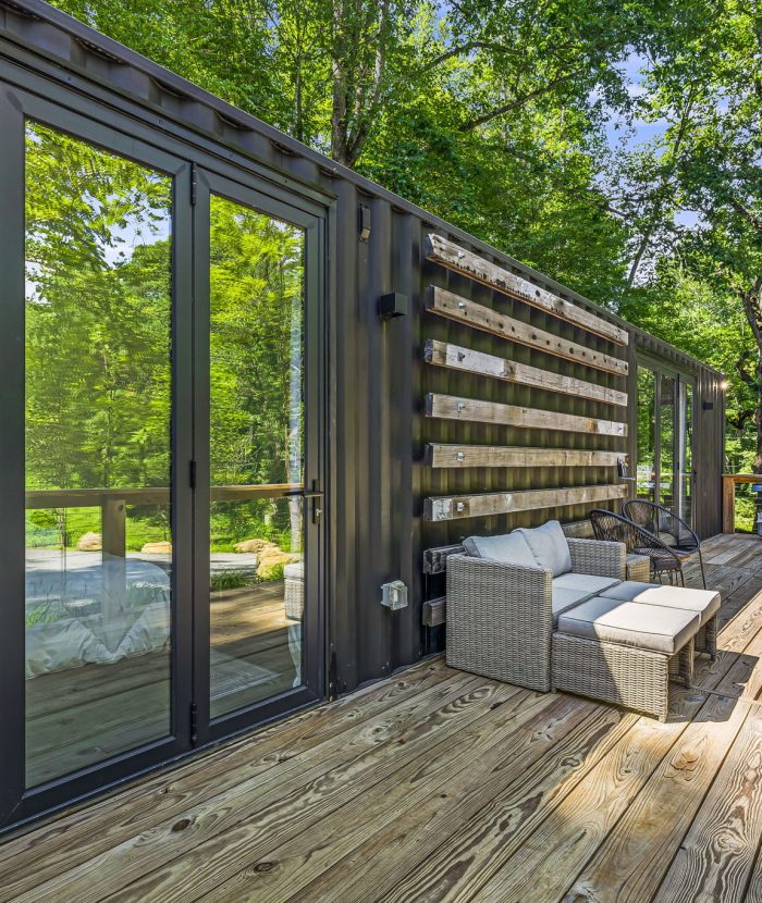 A wooden deck with outdoor seating, including a cushioned chair and sofa. Glass doors reflect a green wooded area. A grill is situated near the railing.