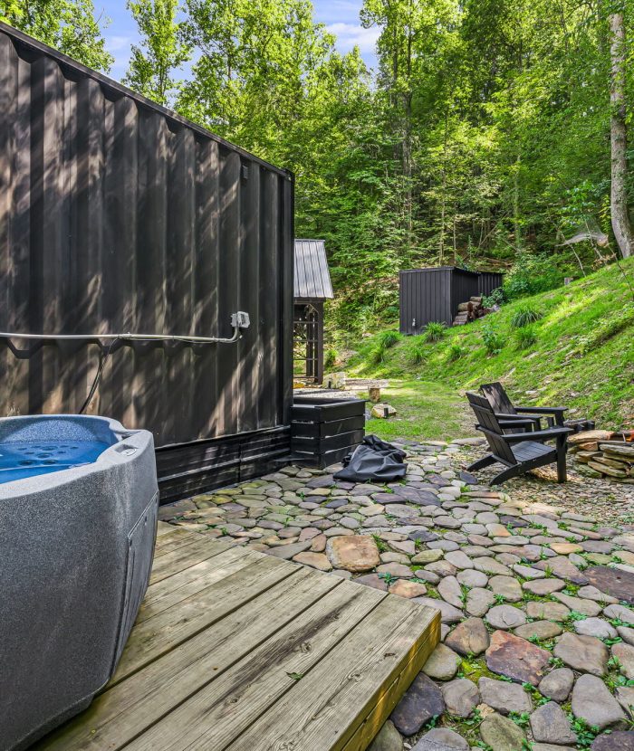 Outdoor area with a hot tub on a wooden deck, a stone-paved patio with Adirondack chairs, a small fire pit, and lush green surroundings.