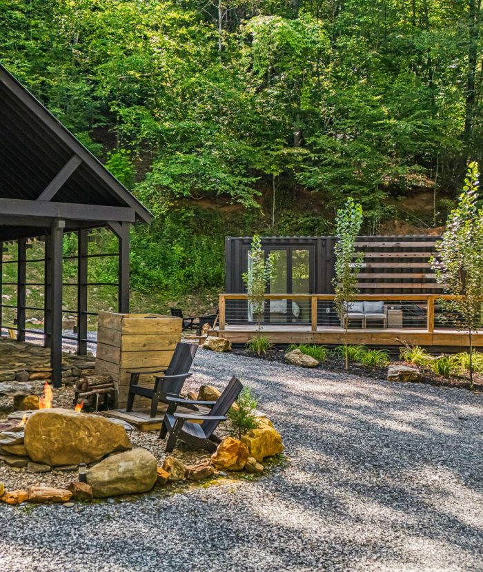 Outdoor area with a covered seating structure on the left and a modern cabin deck on the right, surrounded by trees and greenery with a gravel foundation and a small fire pit in front.