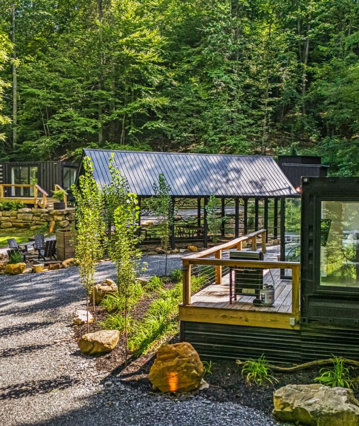 A modern cabin complex in a wooded area featuring black exterior finishes, large windows, deck spaces, gravel pathways, and surrounded by lush greenery.