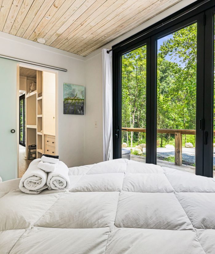 A bedroom with a wooden ceiling, large glass doors opening to a wooded area, a white bed with folded towels, a small bench, and an open door leading to a walk-in closet and desk area.