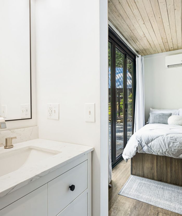 A small, modern bedroom with a neatly made bed, white linens, and an air conditioner. Adjacent is a minimalist bathroom with a white vanity and sink, set against a wooden ceiling backdrop.