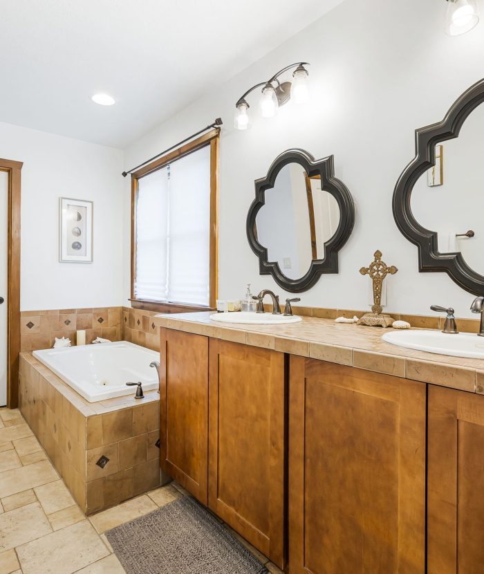 A bathroom with a wooden vanity, dual sinks, ornate mirrors, a jacuzzi tub, and beige tile floors. A white door and a window with white blinds are visible.