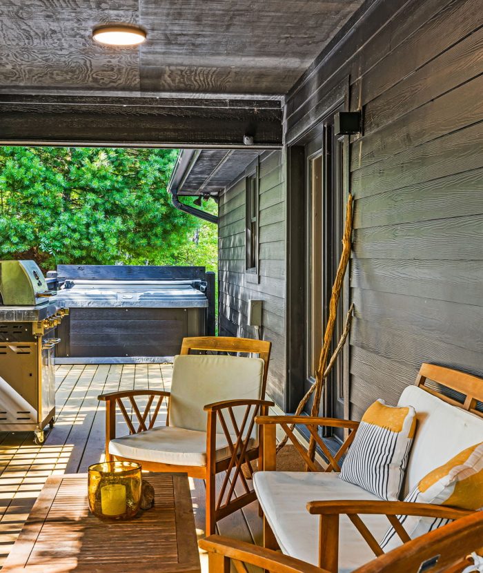 A covered wooden deck features a grill, seating area with wooden chairs and cushions, and natural light streaming in. A lush, green background is visible beyond the wooden railing.