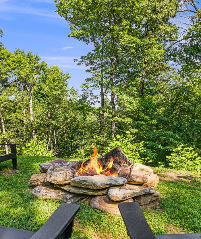 Outdoor campfire with wooden chairs set in a circle on a grassy area surrounded by lush green trees.