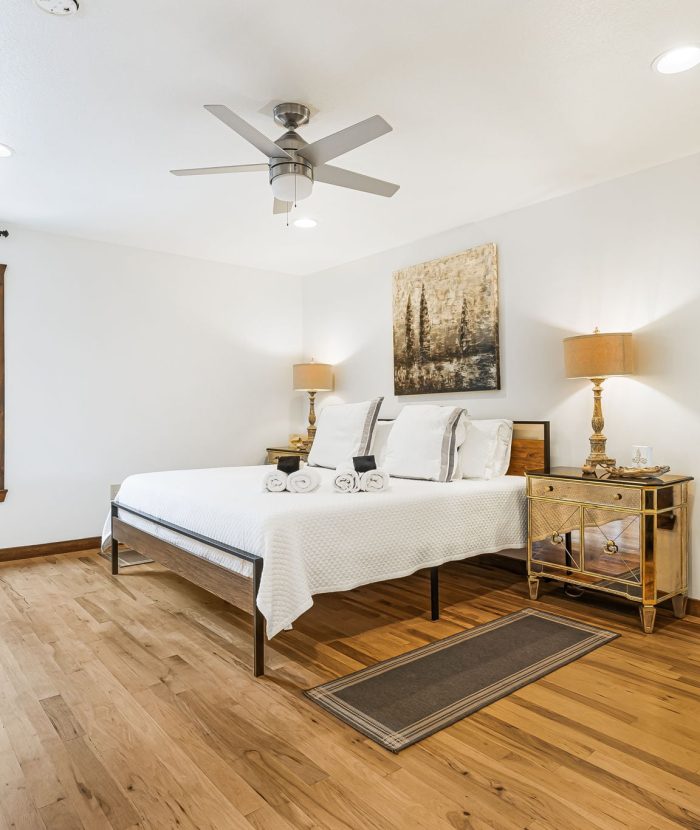 A bedroom with hardwood floors, a bed with white linens, two nightstands with lamps, a ceiling fan, artwork above the bed, large windows with white curtains, and a view of greenery outside.