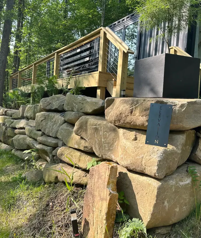 Our Elevation Eco-Villa built with shipping containers, featuring a wooden deck and surrounded by a large stone retaining wall in a forested area.