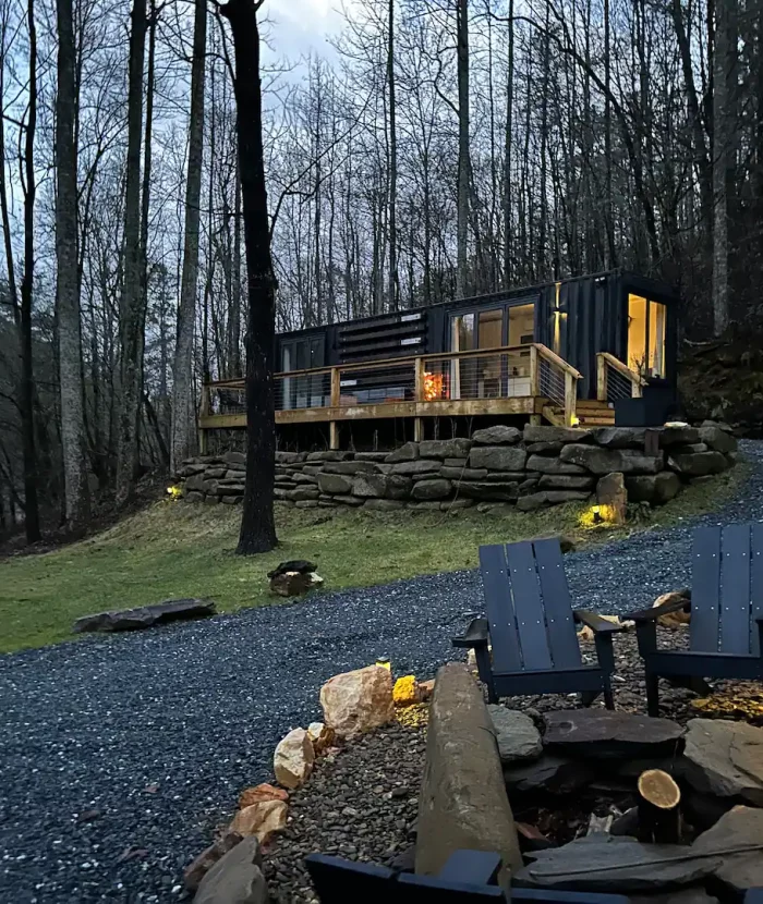 Our Elevation Eco-Villa is a modern cabin with large windows and a deck illuminated at dusk, set in a wooded area, with a foreground featuring a fire pit and two adirondack chairs.