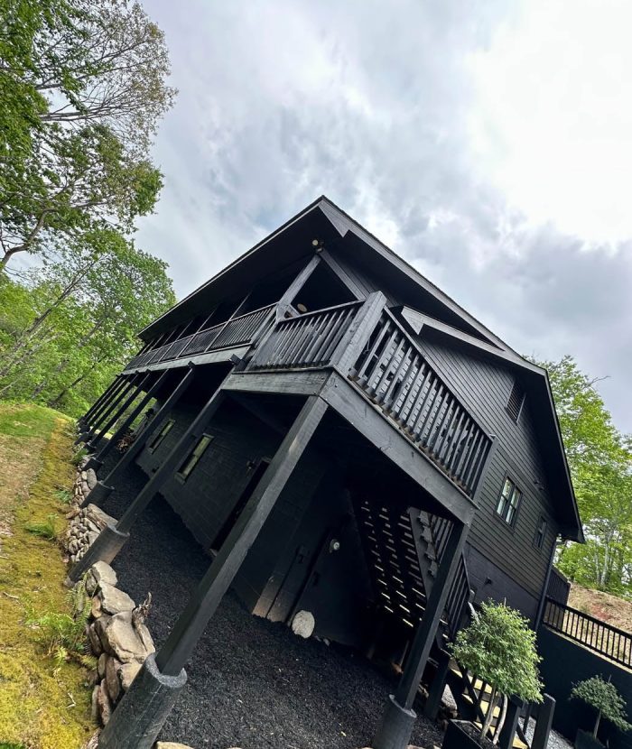 A black wooden house with a wrap-around porch and elevated foundation, surrounded by trees and greenery under an overcast sky. A staircase leads down from the porch to the ground below.